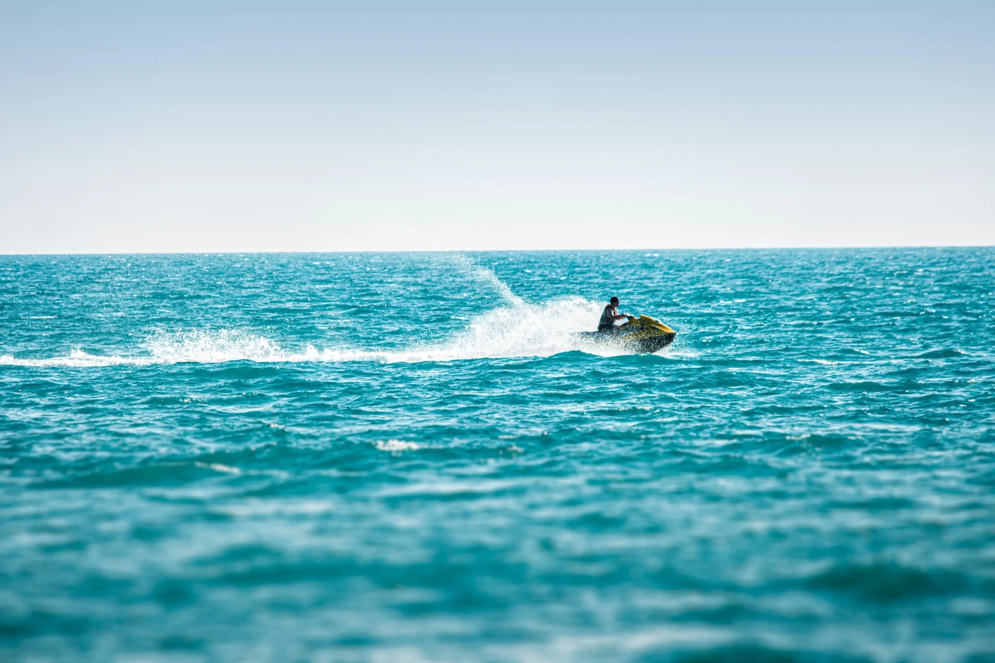 Person riding a jet ski on a clear blue ocean with a light blue sky.